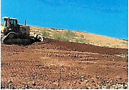 A bulldozer sits on the crest of a bare earthen slope at the landfill under a clear sky, with loose reddish-brown soil and scattered stone covering the foreground.